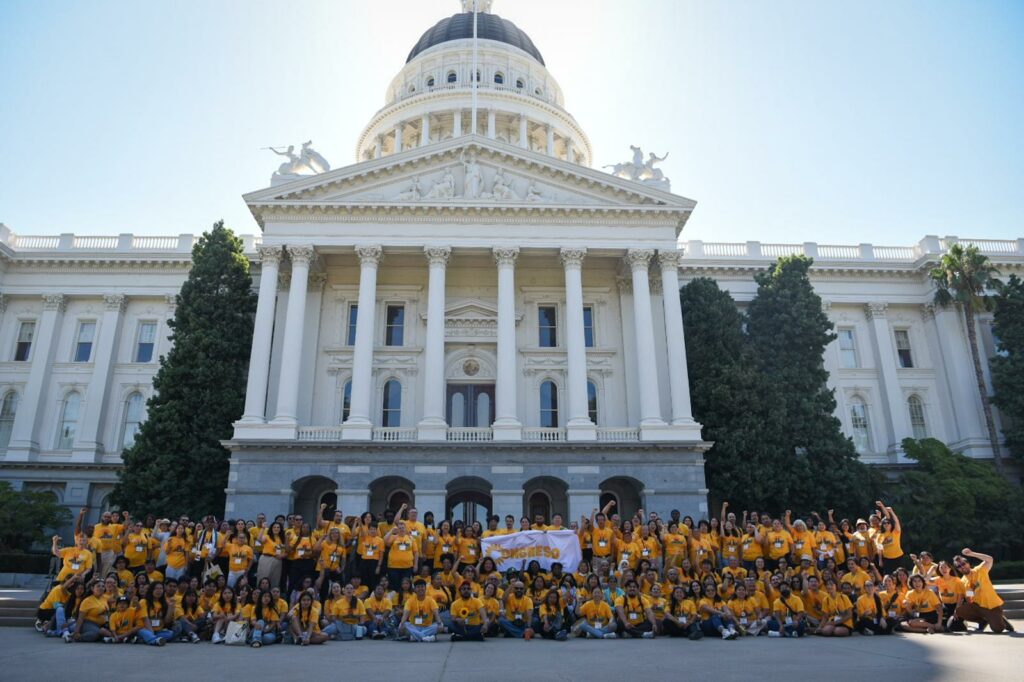 A large group of people in EHC yellow shirts poses in front of a classical white capitol building with pillars and a dome, holding a white banner. Photo by Brooke Anderson.